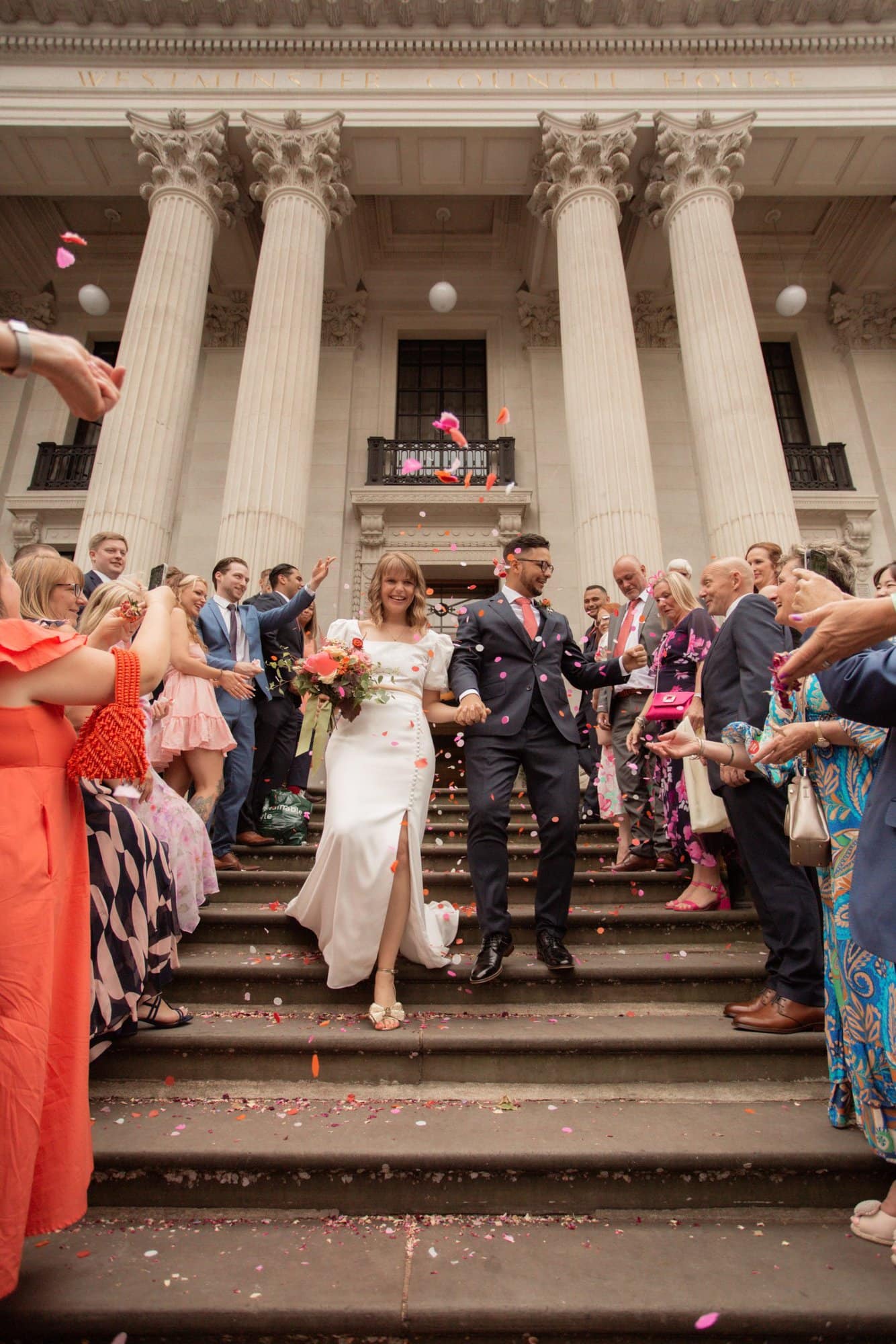 Newlywed couple being showered with confetti whilst walking down a tunnel of their friends and family down the steps of Marylebone Town Hall Registry office after their Wedding in London